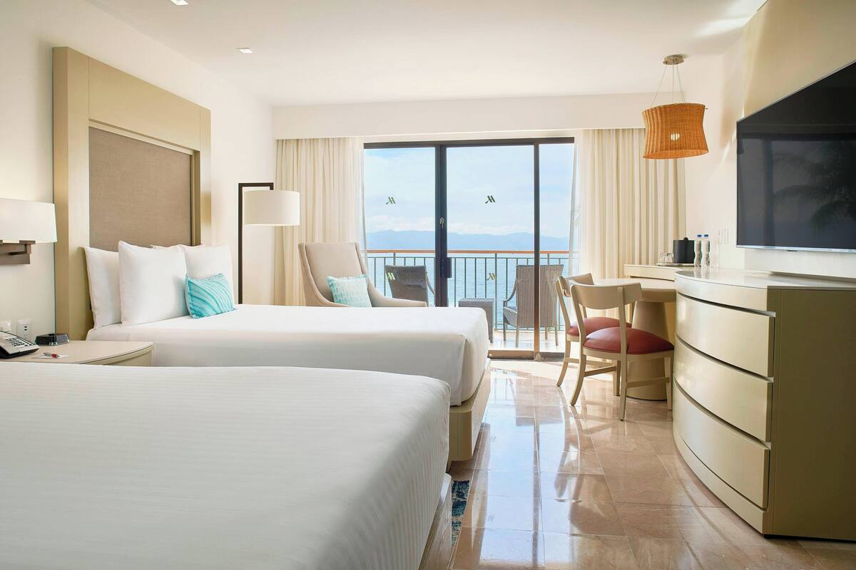 A hotel room with two beds, a desk, a TV, and a balcony with a view.