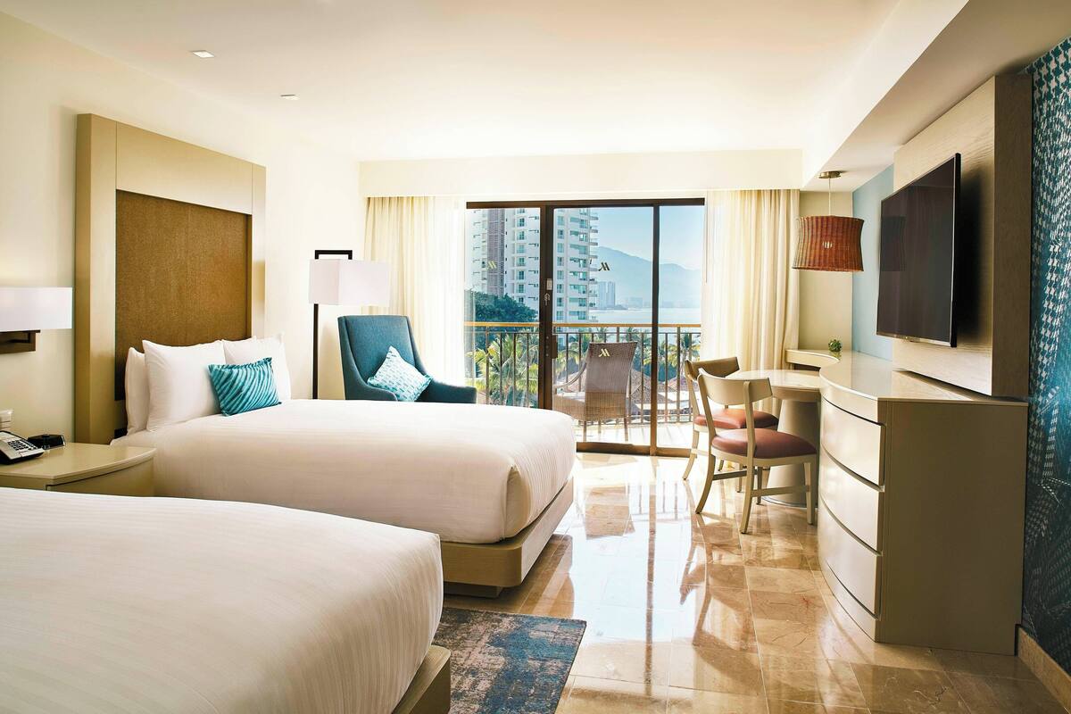 A hotel room with two beds, a desk, a TV, and a balcony with a view of buildings.