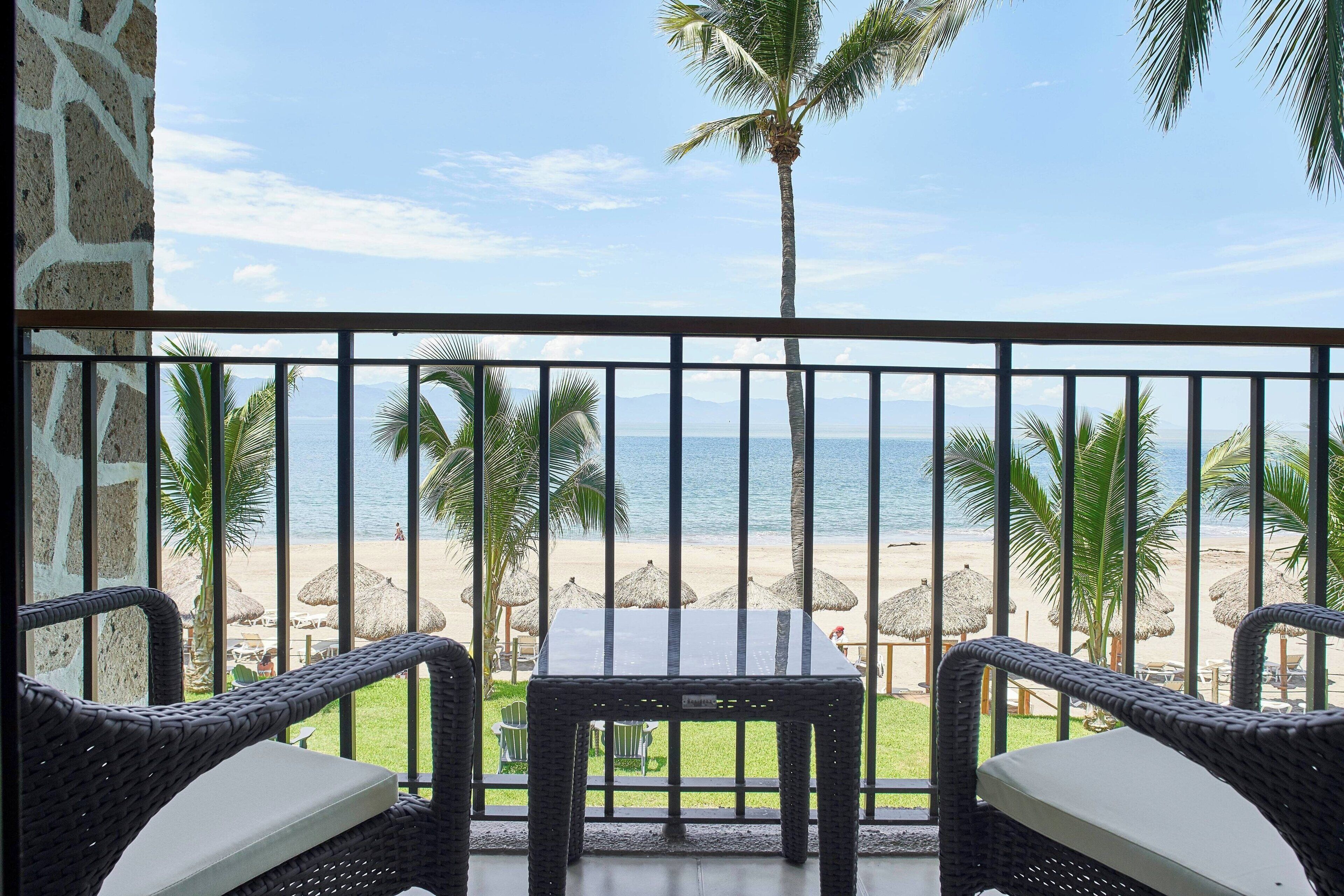 A balcony with wicker furniture overlooking a beach with palm trees and thatched umbrellas.