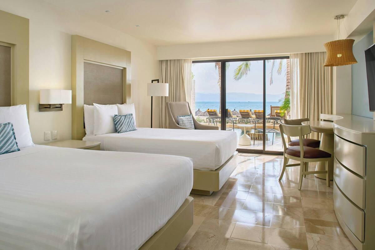 A hotel room with two beds, a desk, a chair, and a view of the ocean.