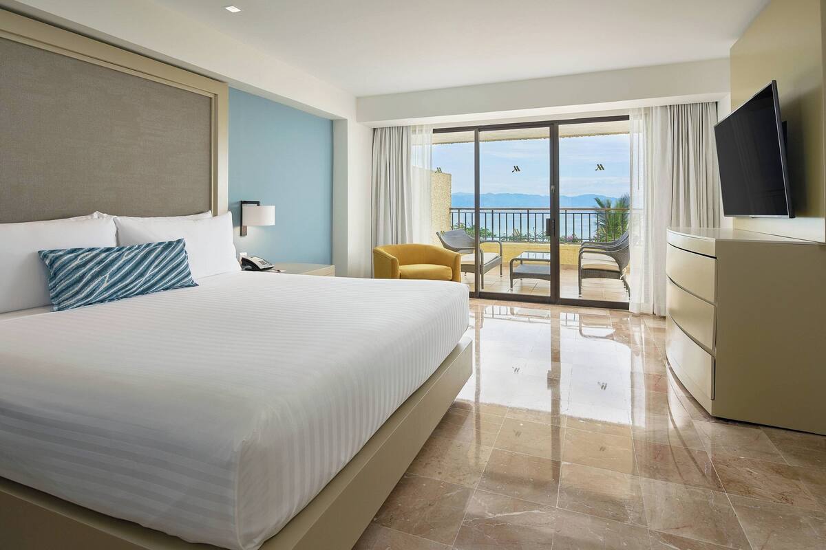 A hotel room with a large bed, a TV, a desk, and a balcony with a view of the mountains.