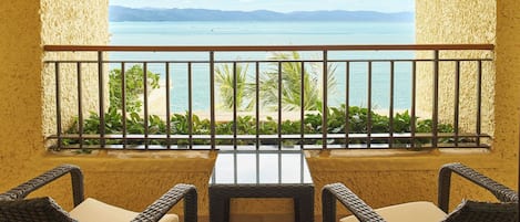 A balcony with wicker chairs and a table, overlooking a beach and the sea.