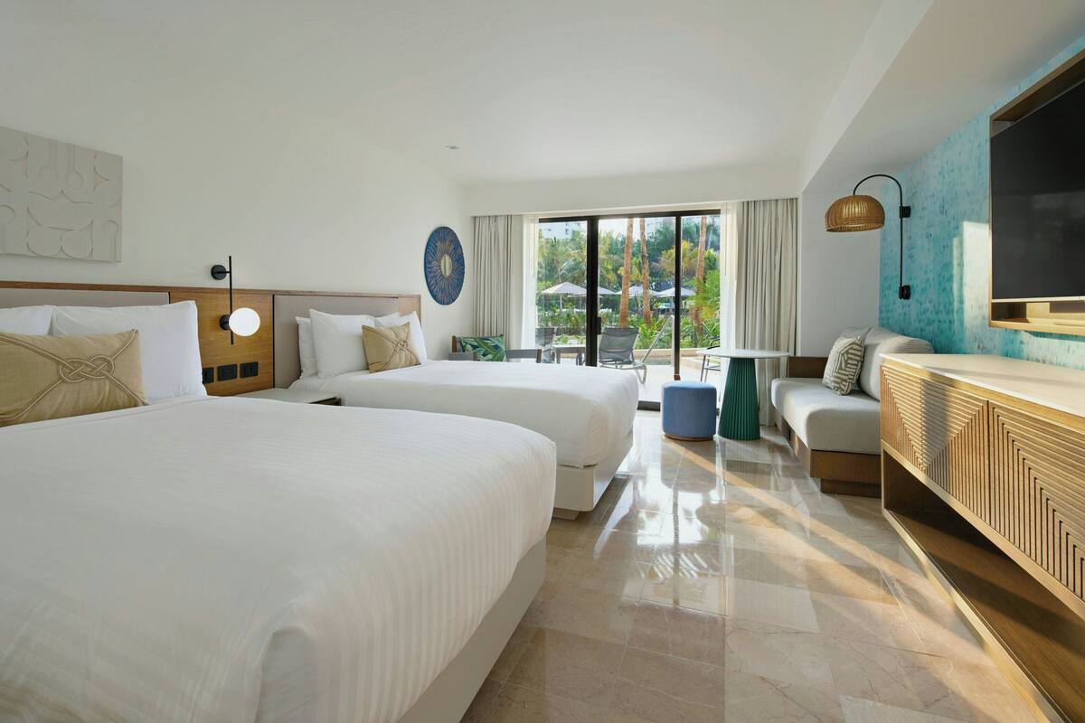 A hotel room with two large beds, a sofa, a TV, and a balcony with a view of palm trees.