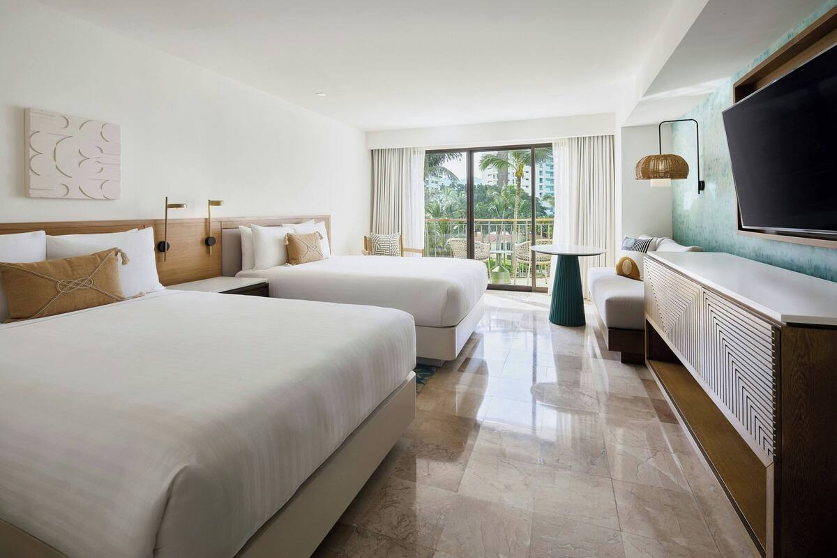 A hotel room with two beds, a balcony with a table and chairs, a TV, and a large window with a view of palm trees.