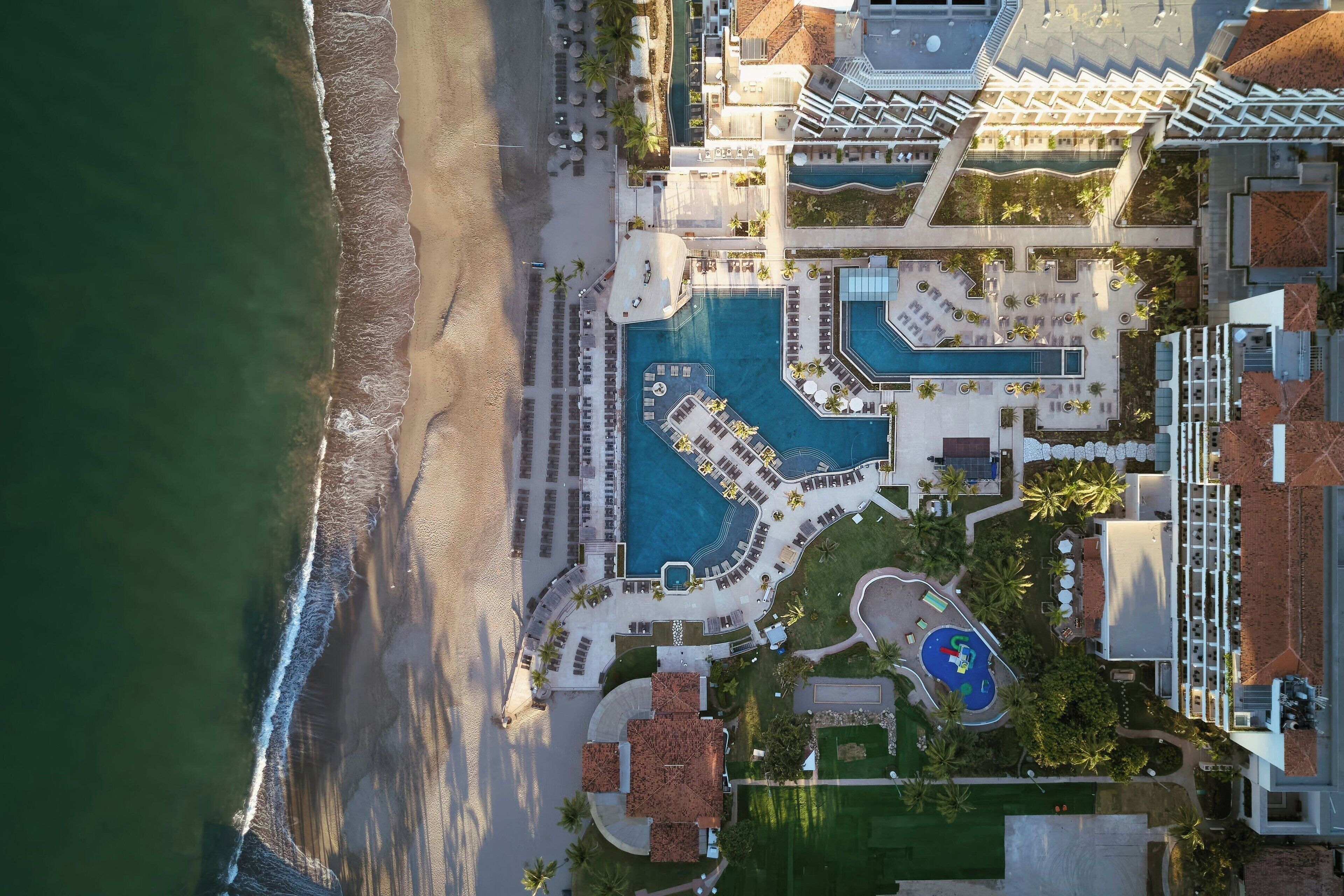 An aerial view of a resort with a large pool, beach access, and multiple lounge areas.