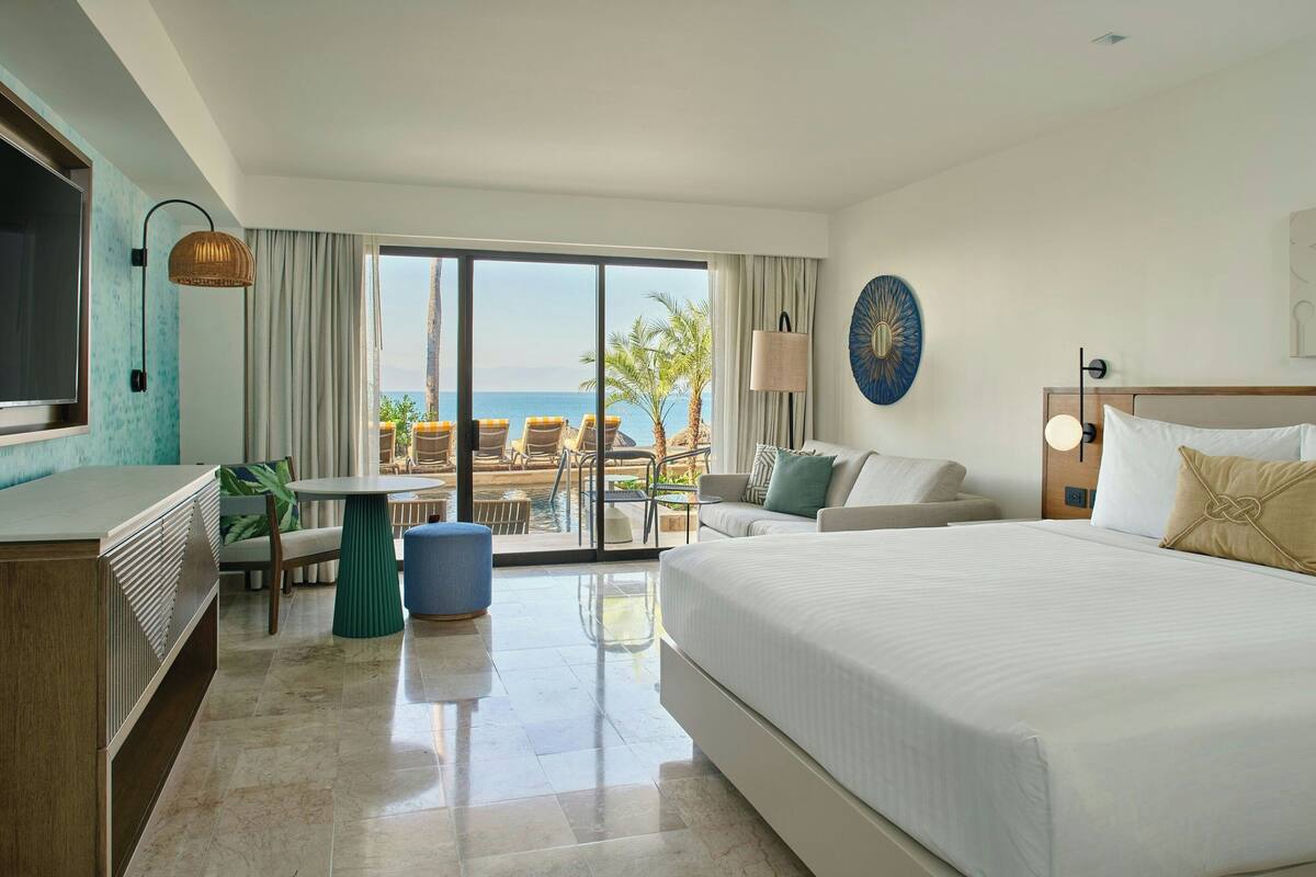 A hotel room with a large bed, a TV, a dining table, and a view of the ocean.