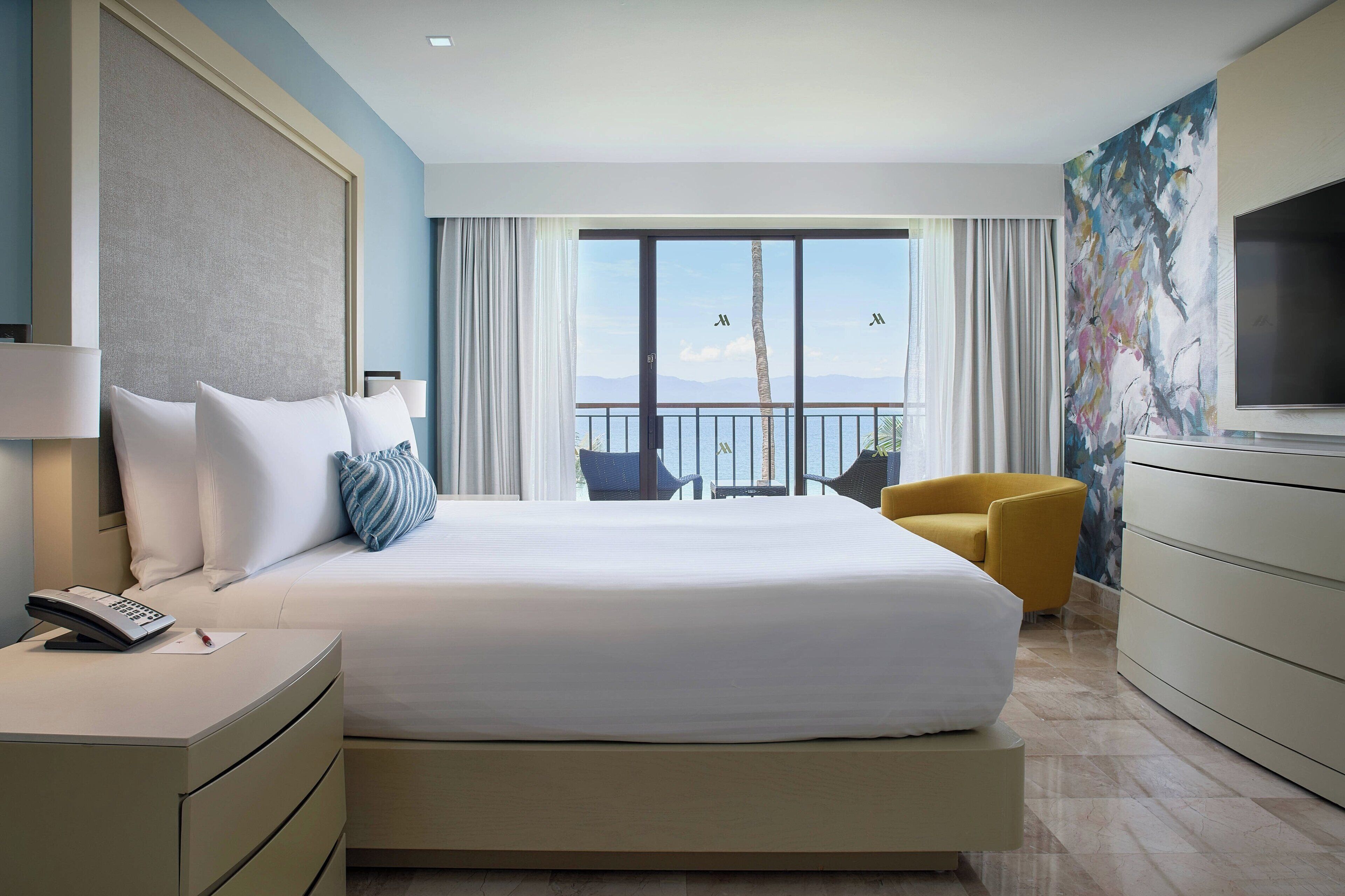 A hotel room with a large bed, a desk with a telephone, a chair, a television, and a balcony with a view of the sea.