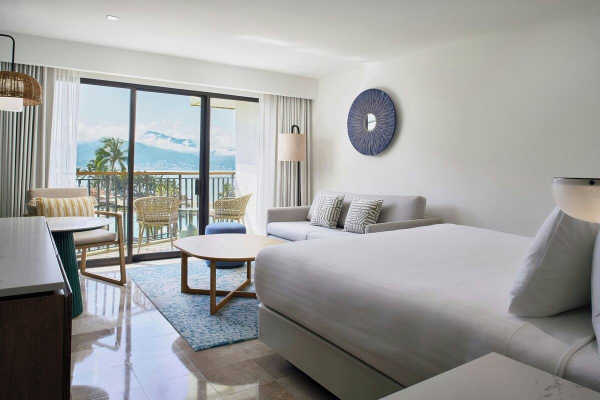 A hotel room with a large bed, a sofa, a dining table, and a balcony with a view of mountains.