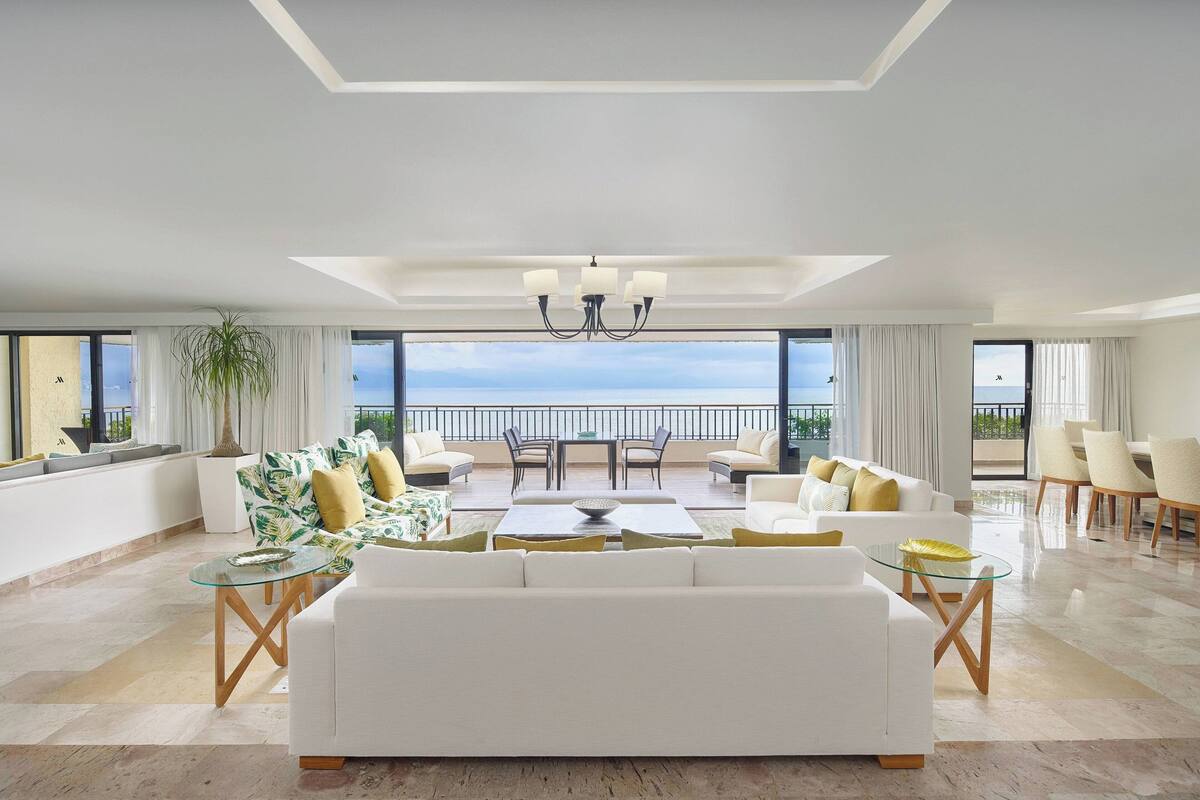 A spacious living room with a large white sofa, a glass coffee table, and a dining area with chairs and a table. A large window offers a view of the sea.