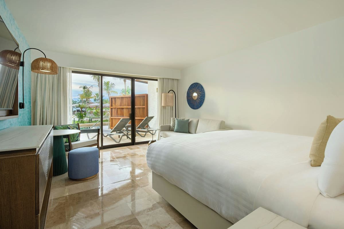 A hotel room with a large bed, a desk, a chair, a small table, and a view of the outdoors.