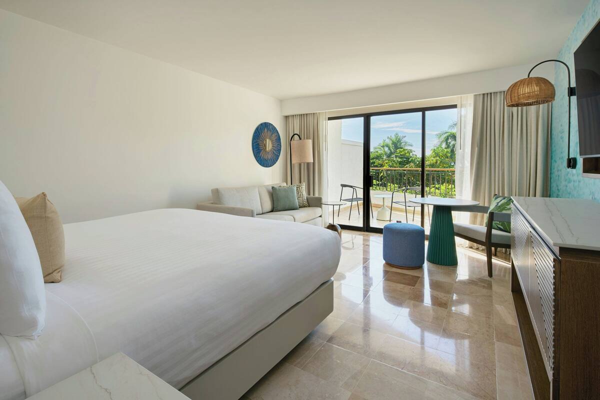 A hotel room with a large bed, a sofa, a dining table, and a view of the outdoors.