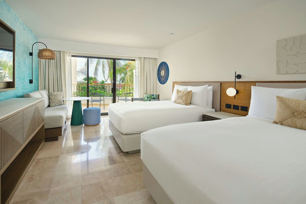 A hotel room with two beds, a balcony with a view of palm trees, a dining area, and a large window.