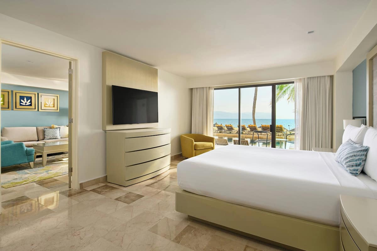 A hotel room with a large bed, a TV mounted on the wall, a view of the sea, and a balcony with outdoor seating.