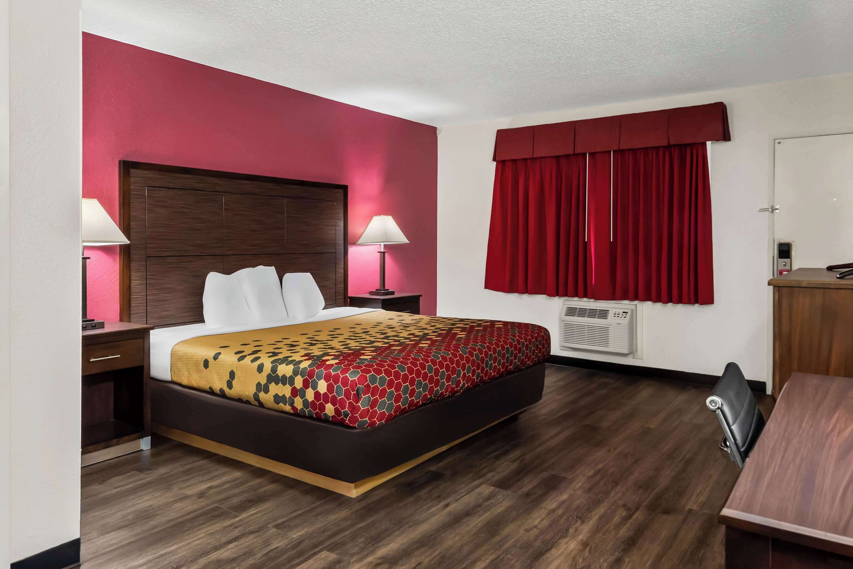 A hotel room with a bed, bedside tables, a desk, and a window with red curtains.