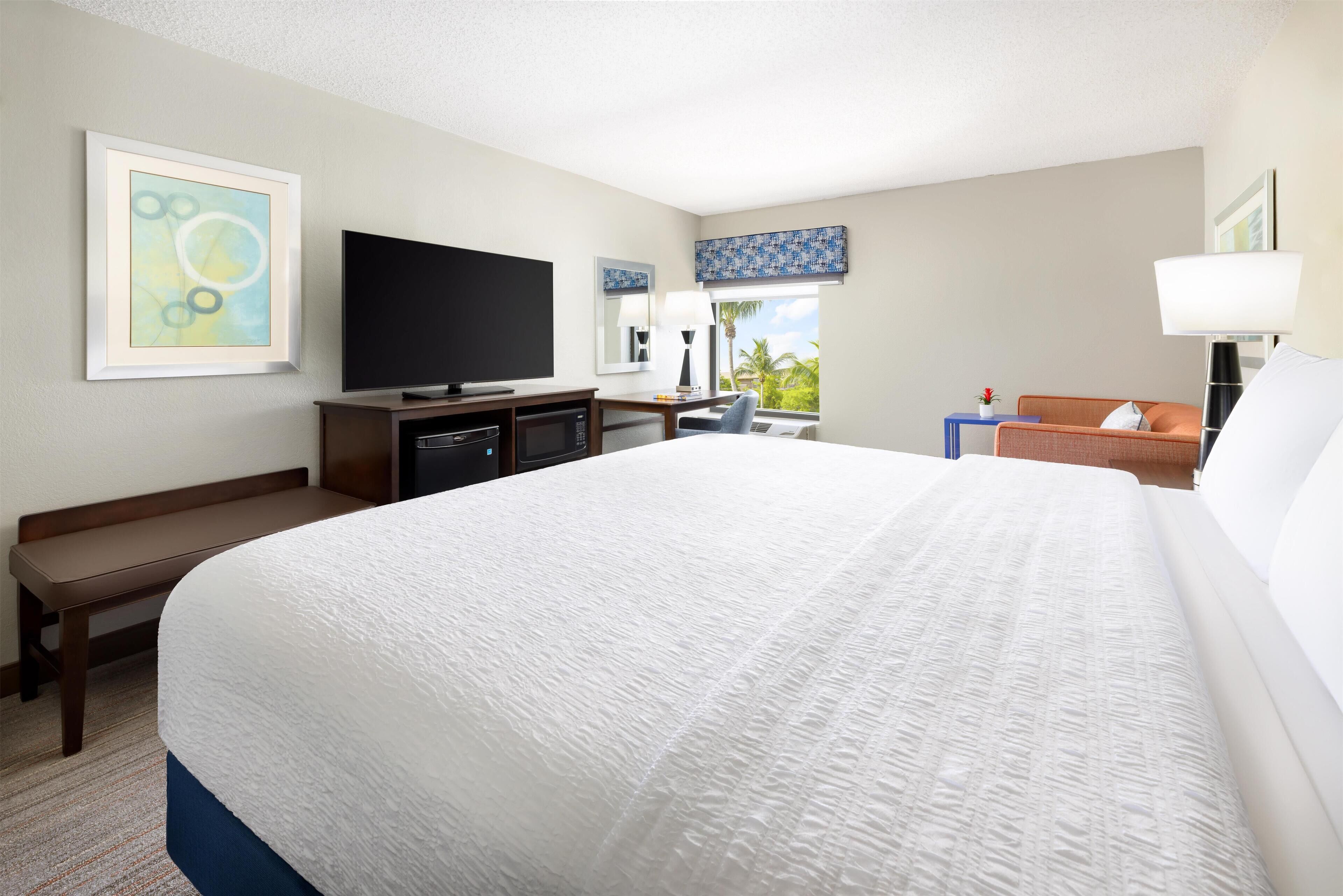 A hotel room with a large bed, a flat-screen TV, a desk with a computer, a chair, and a window with a view of palm trees.