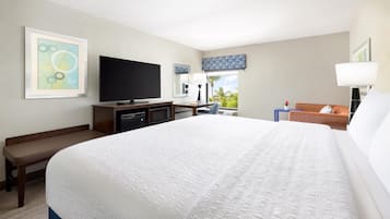 A hotel room with a large bed, a flat-screen TV, a desk with a computer, a chair, and a window with a view of palm trees.