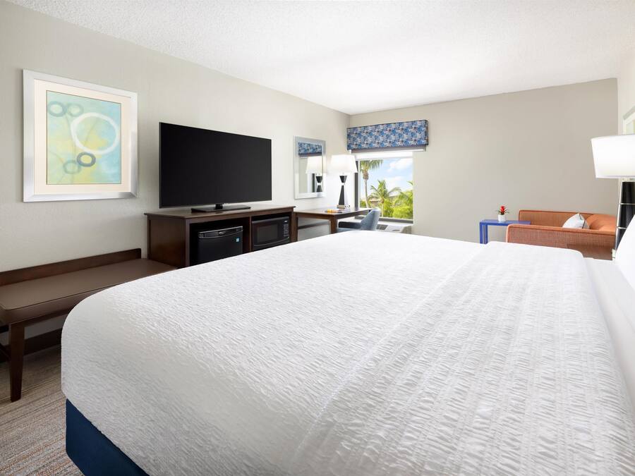 A hotel room with a large bed, a flat-screen TV, a desk with a computer, a chair, and a window with a view of palm trees.
