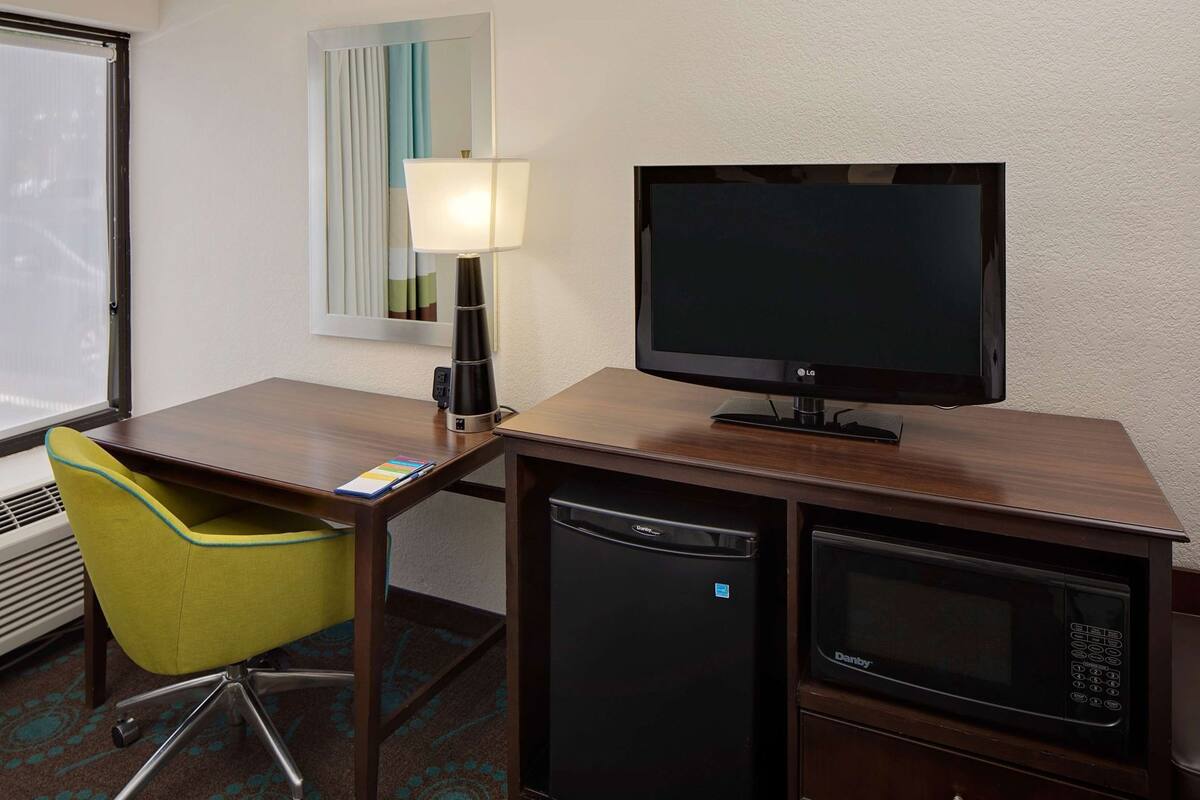 A hotel room with a wooden desk, a green chair, a black TV, a lamp, a mini-fridge, and a microwave.