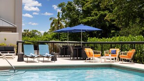 A poolside area with lounge chairs and a table, surrounded by a fence and greenery.