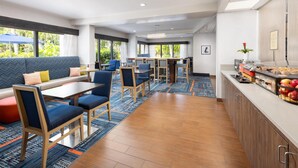 A modern hotel lobby with a seating area, a dining table, and a kitchen counter with food items.