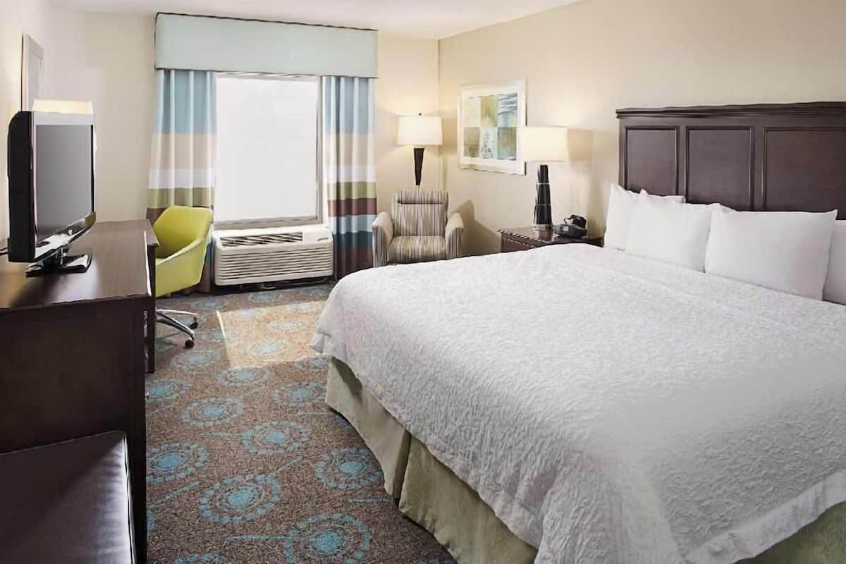 A hotel room with a large bed, a desk with a computer, a chair, a lamp, and a window with curtains.