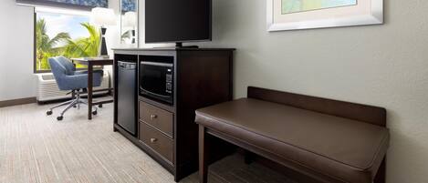 A hotel room with a flat-screen TV, a desk with a chair, a bench, a microwave, and a large window with a view of palm trees.