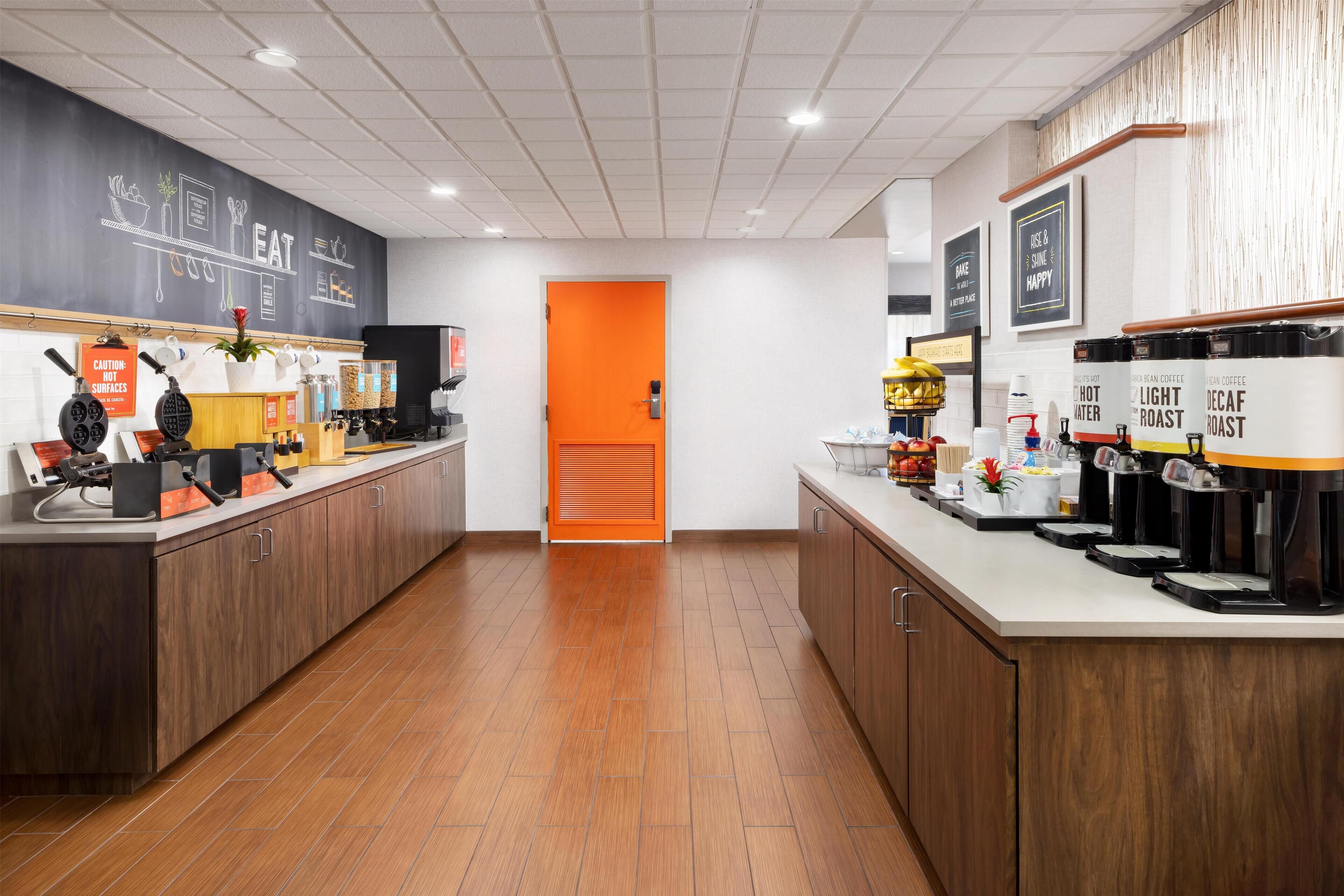 Une cuisine moderne avec des meubles en bois, un tableau noir orné de dessins sur le thème de la nourriture et une porte d’un orange vif.