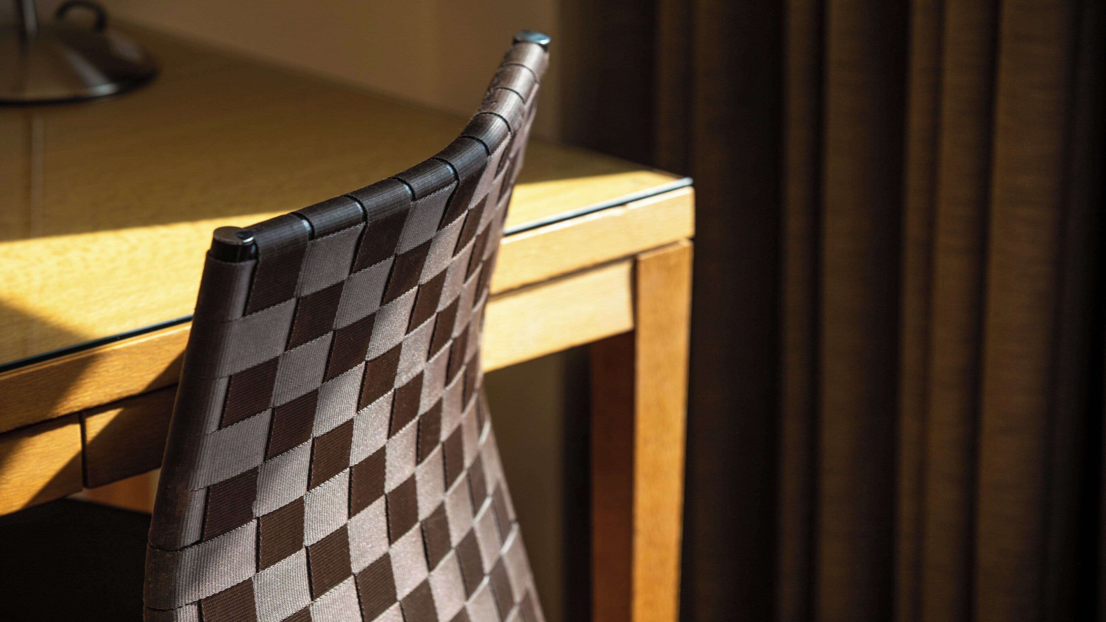 A chair with a checkered pattern, positioned at a wooden table with a lamp in the background.