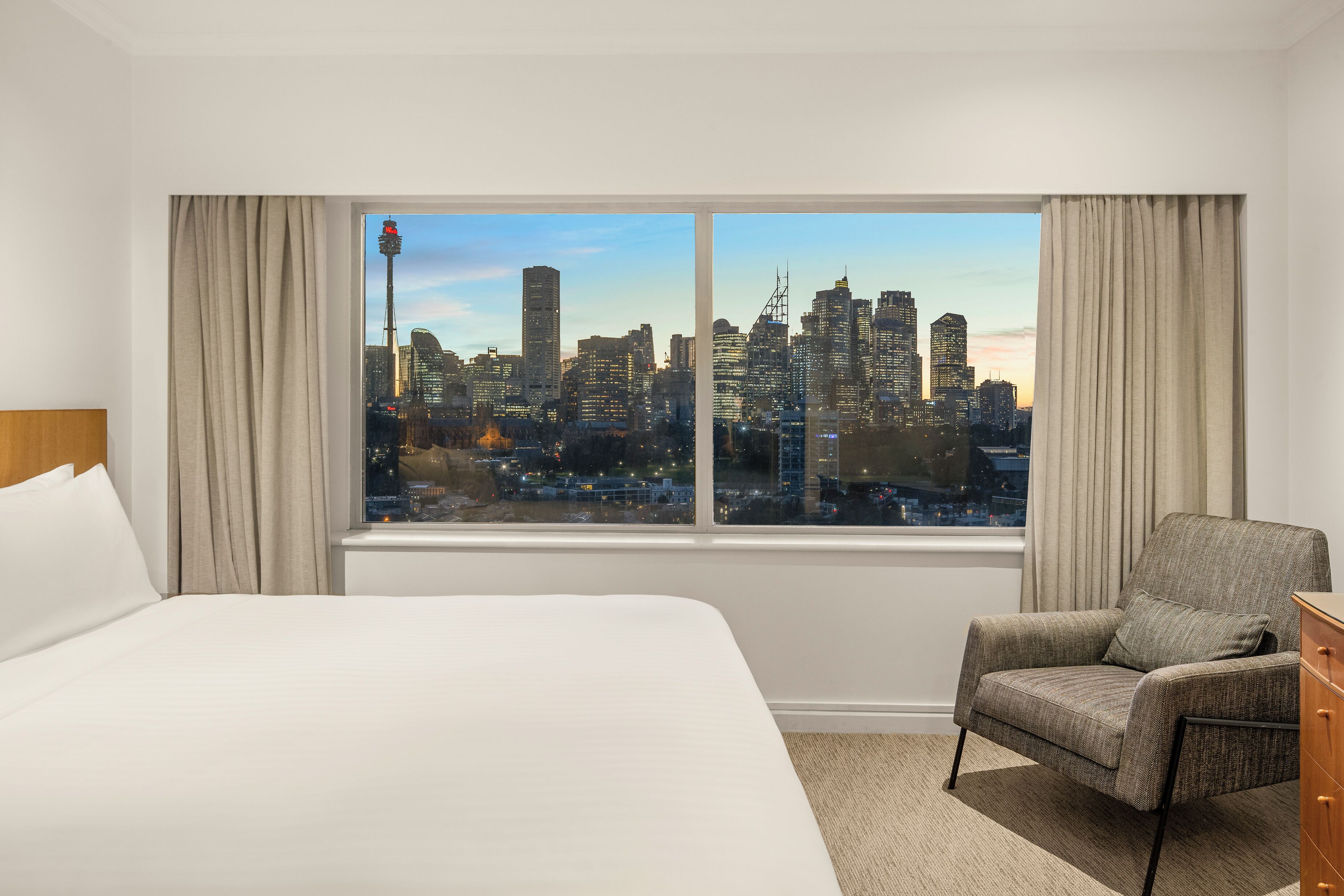 A hotel room with a large bed, a chair, a dresser, and a window offering a city skyline view.
