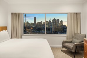 Habitación de hotel con una cama grande, una silla, un aparador y una ventana con vistas al horizonte de la ciudad.