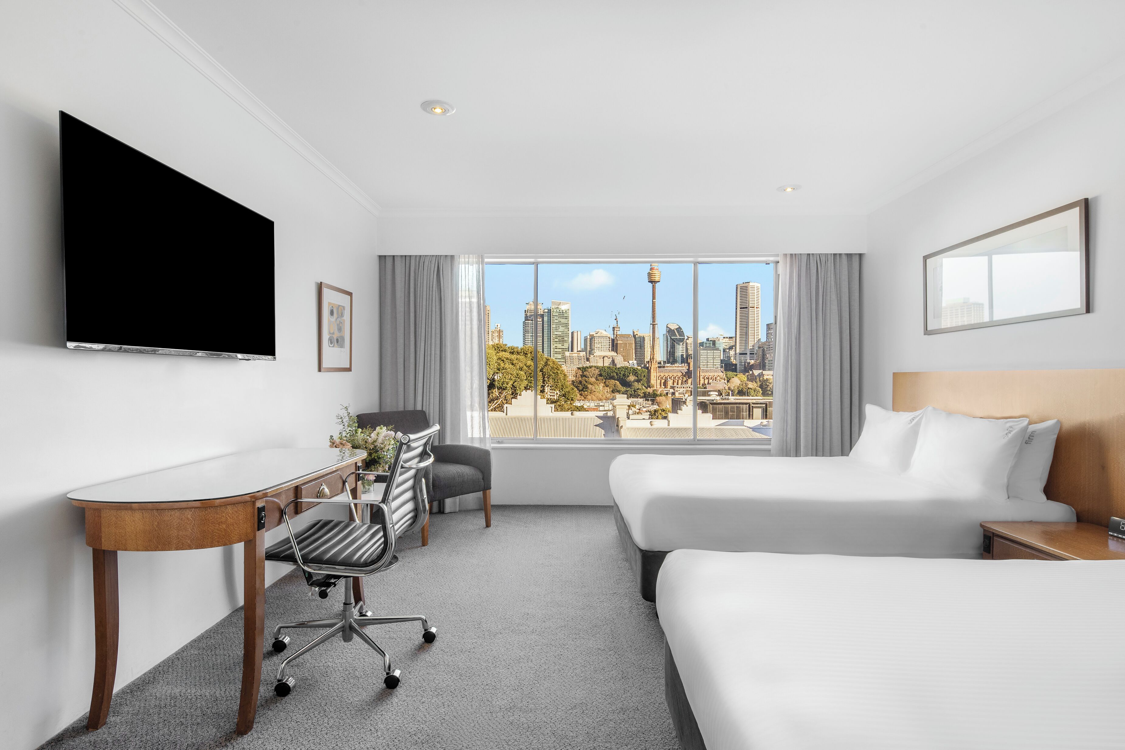 A hotel room with a bed, a desk, a chair, a television, and a large window offering a city view.