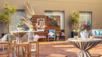 Un patio al aire libre con mobiliario de madera, una pared decorativa con un cartel que dice 'Live Take a Selfie' y varias plantas en macetas.