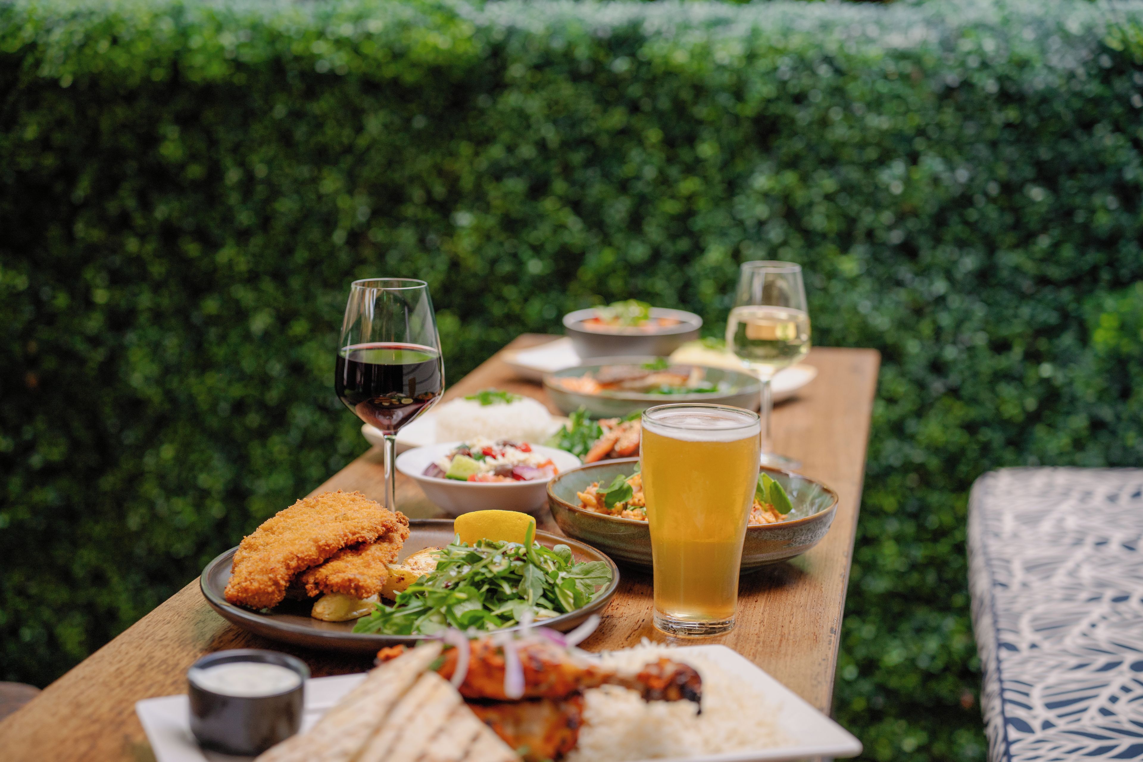 A dining table with various dishes and drinks, set against a green hedge backdrop.