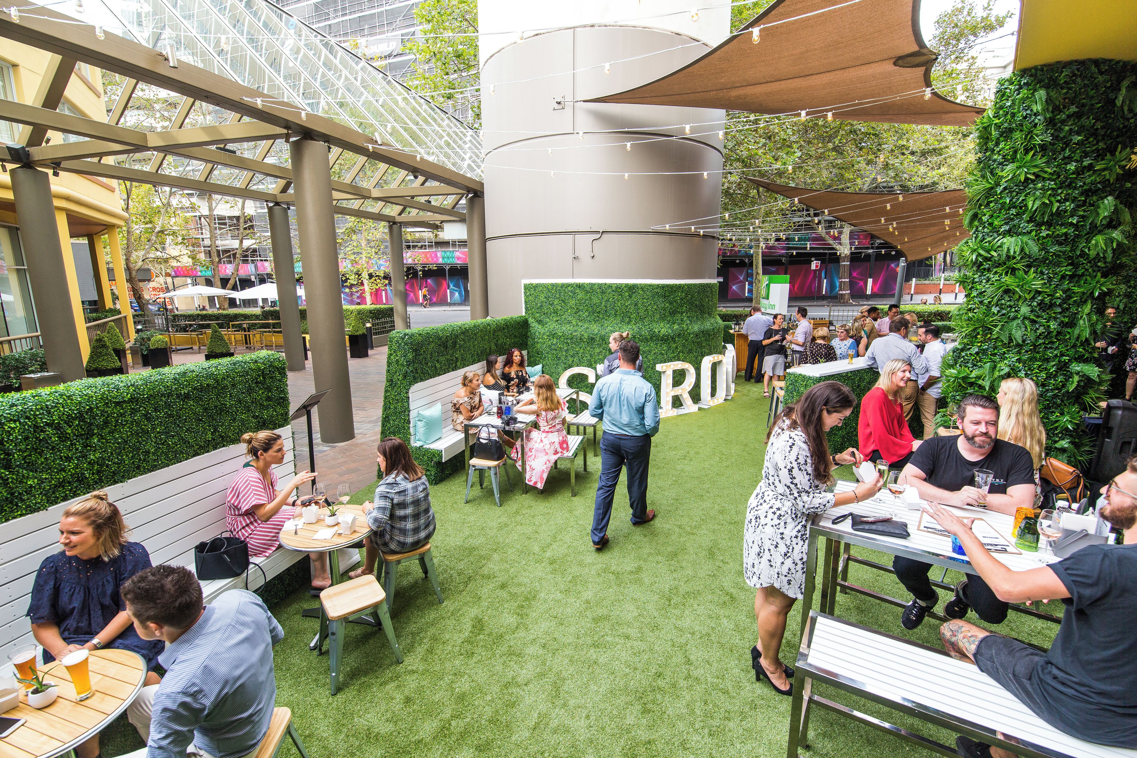 An outdoor seating area with tables and chairs, surrounded by greenery and a modern building structure.