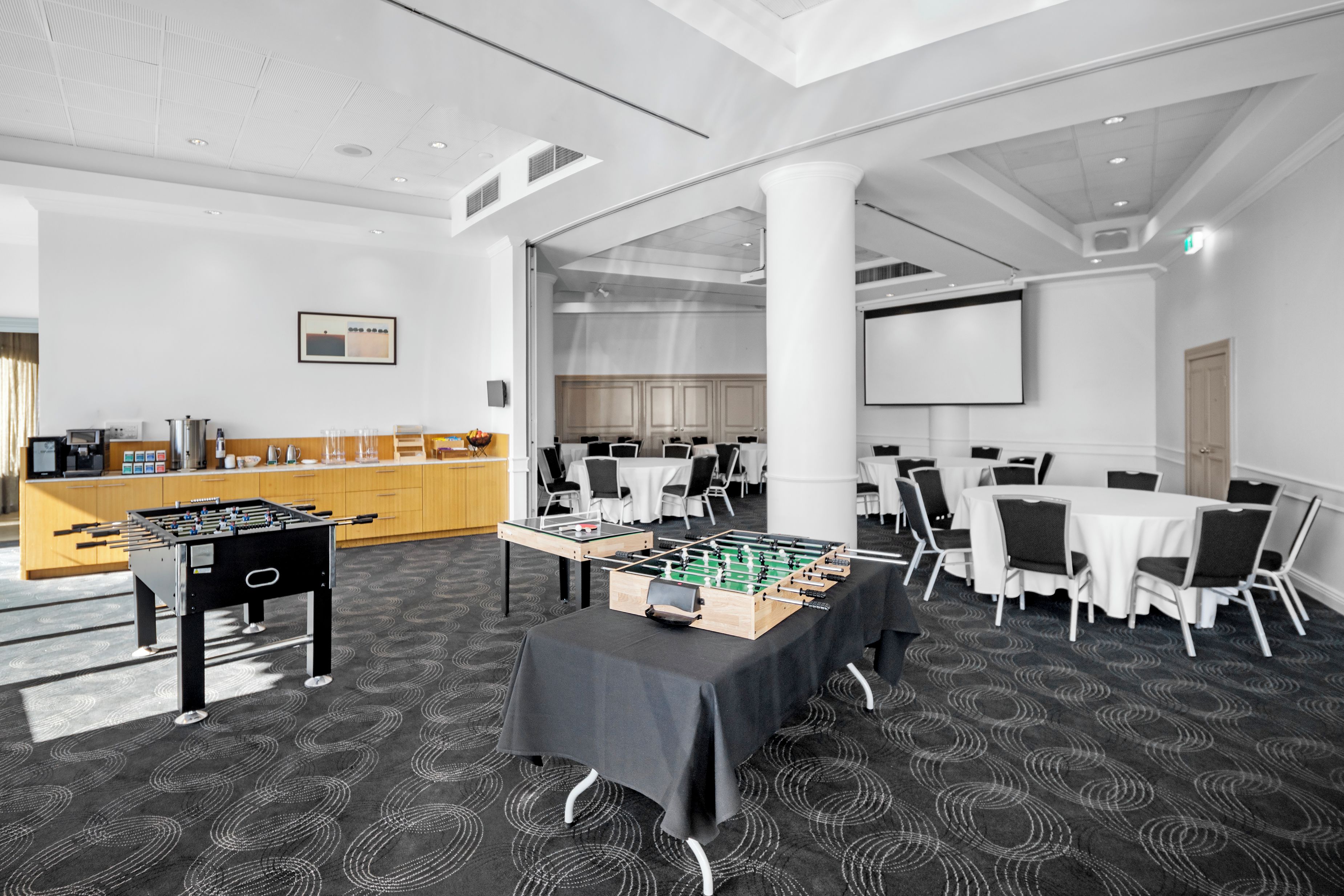 A modern conference room with tables, chairs, a foosball table, and a kitchen area.