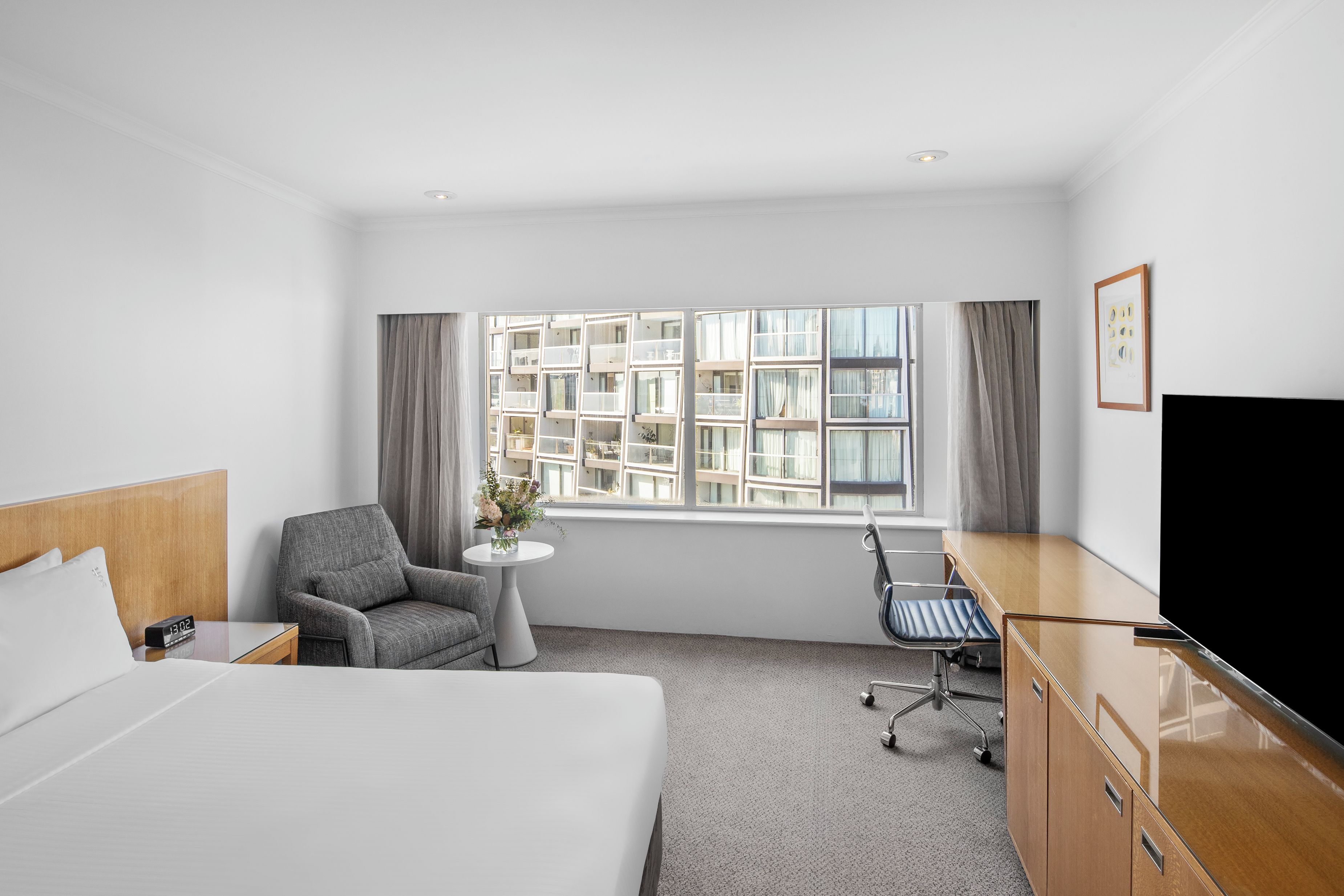 Habitación de hotel con cama, escritorio, silla, televisión y ventana con vista a edificios.