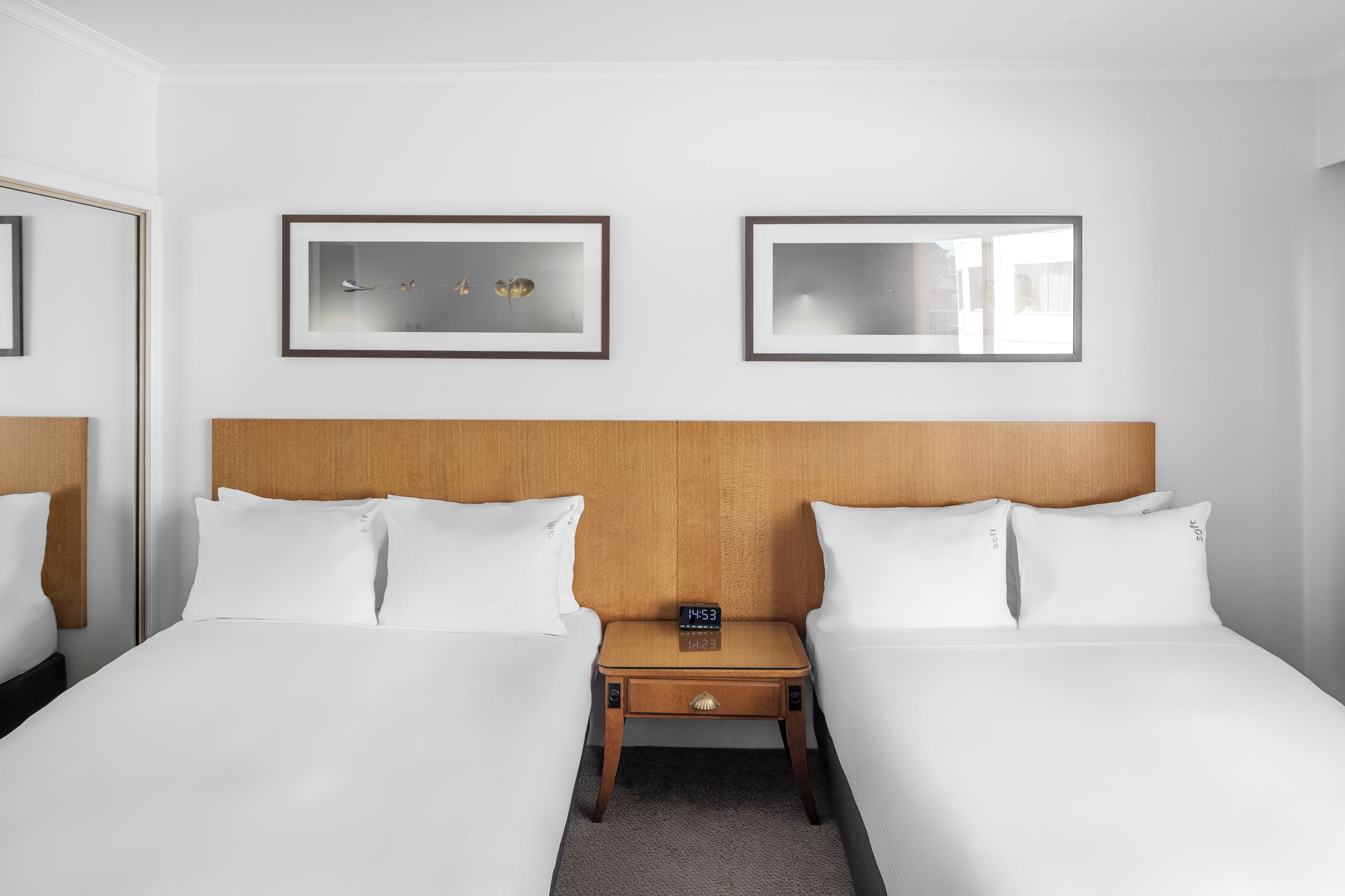 Habitación de hotel con dos camas, un cabecero de madera, una mesita de noche con un reloj y dos cuadros en la pared.