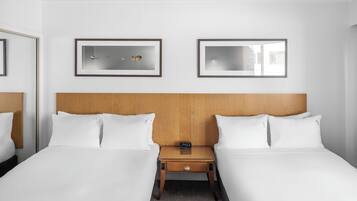 Habitación de hotel con dos camas, un cabecero de madera, una mesita de noche con un reloj y dos cuadros en la pared.