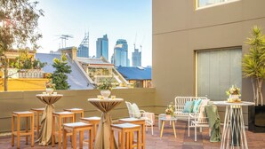 An outdoor patio with wooden furniture, a city skyline view, and a building with a sloped roof. - Holiday Inn Sydney Potts Point by IHG (Potts Point)