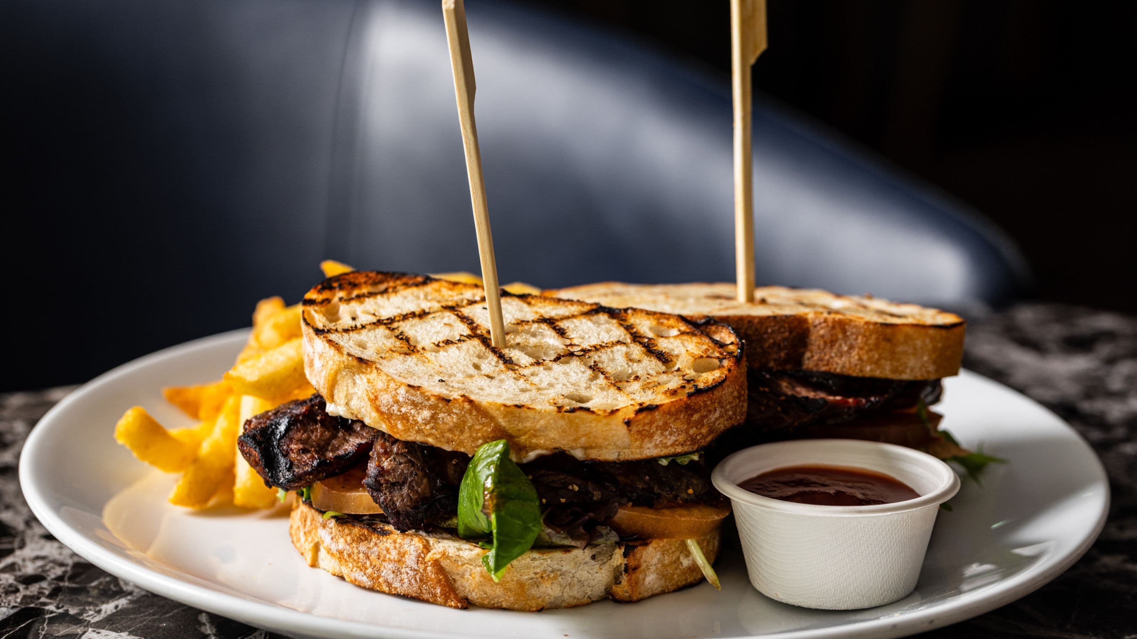 Un sandwich grillé, accompagné d’une portion de frites et d’un petit récipient de sauce, le tout servi sur une assiette.