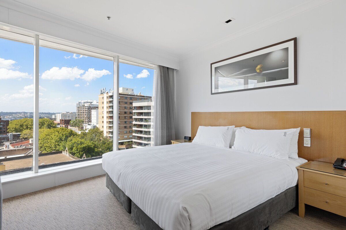 Ein Hotelzimmer mit einem großen Bett, einem Holzkopfstück, einem Nachttisch, einem gerahmten Bild an der Wand und Blick auf Gebäude und Bäume durch ein großes Fenster.