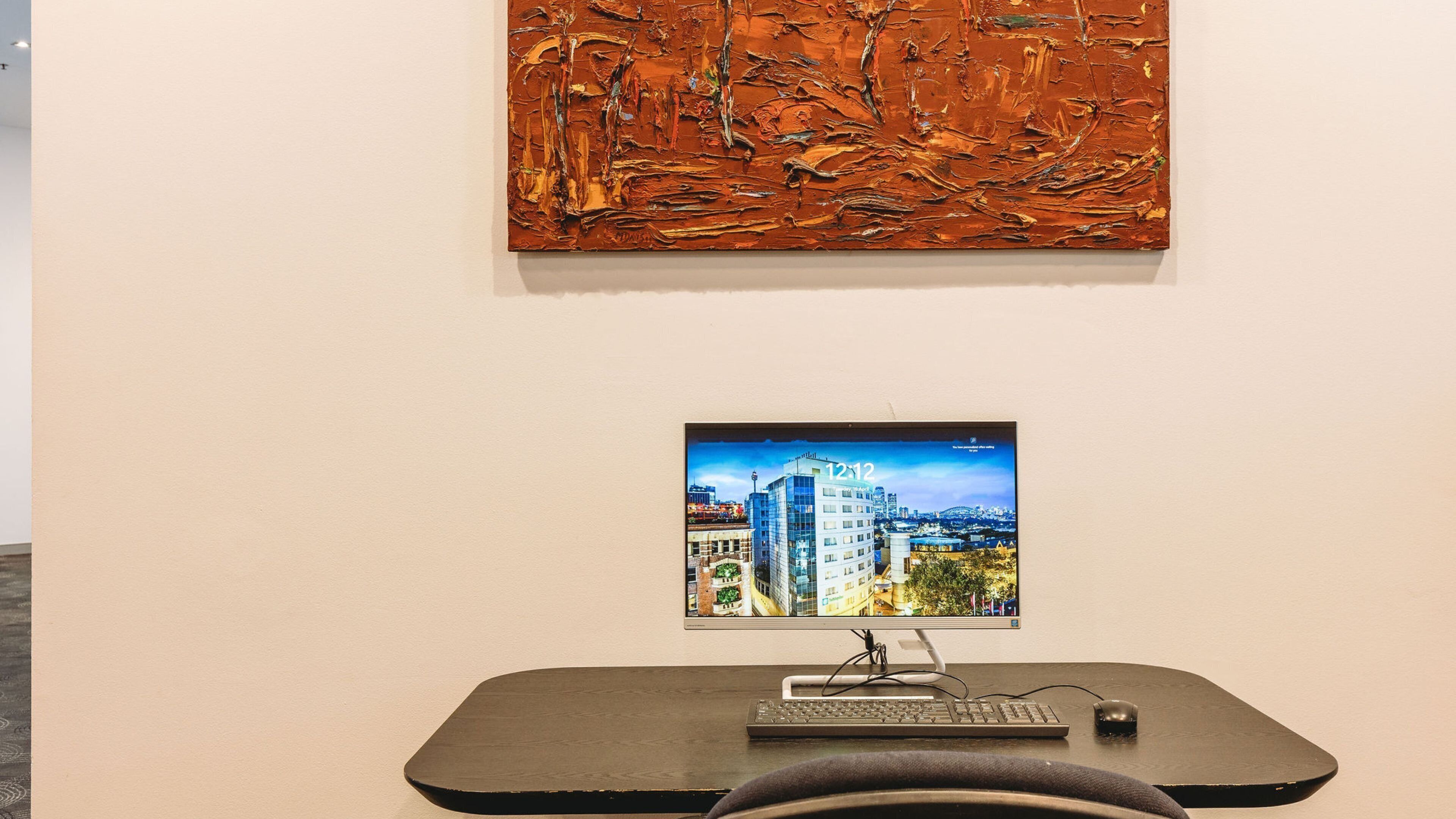 Een scherm op een bureau met toetsenbord en muis, een schilderij erboven en een stoel ervoor.