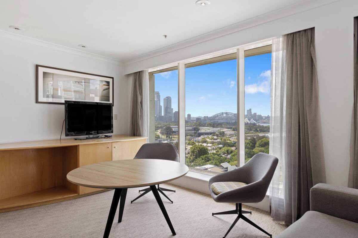 A modern hotel room with a large window offering a city view, a round table, two armchairs, and a flat-screen TV on a wooden console.