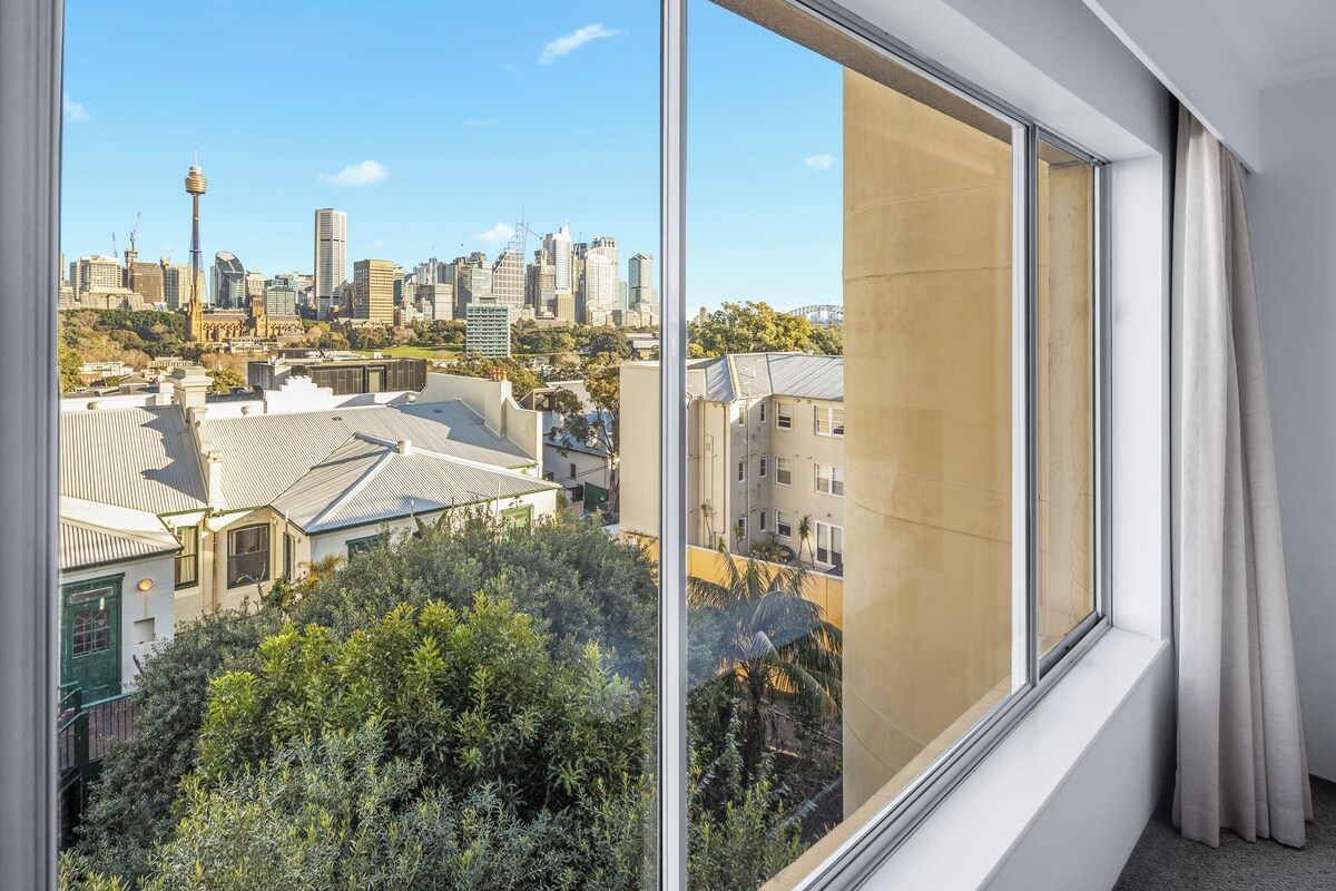 A room with a large window offering a view of a cityscape with various buildings and a hill with greenery.