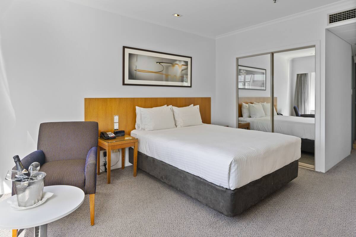 A hotel room with a large bed, a chair, a small table with a drink, and a mirror.