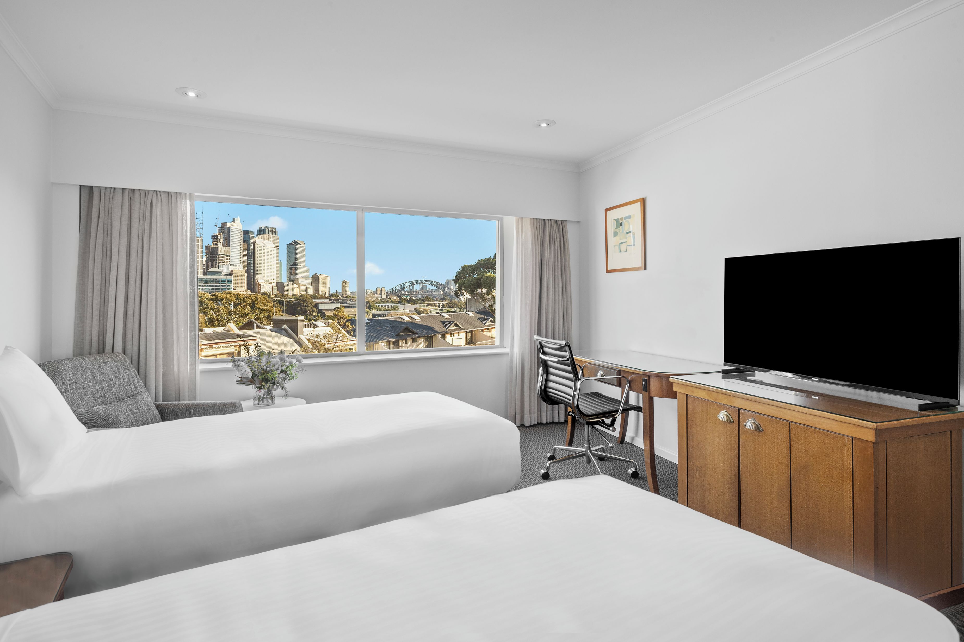 A hotel room with two beds, a desk, a television, and a large window offering a city view.