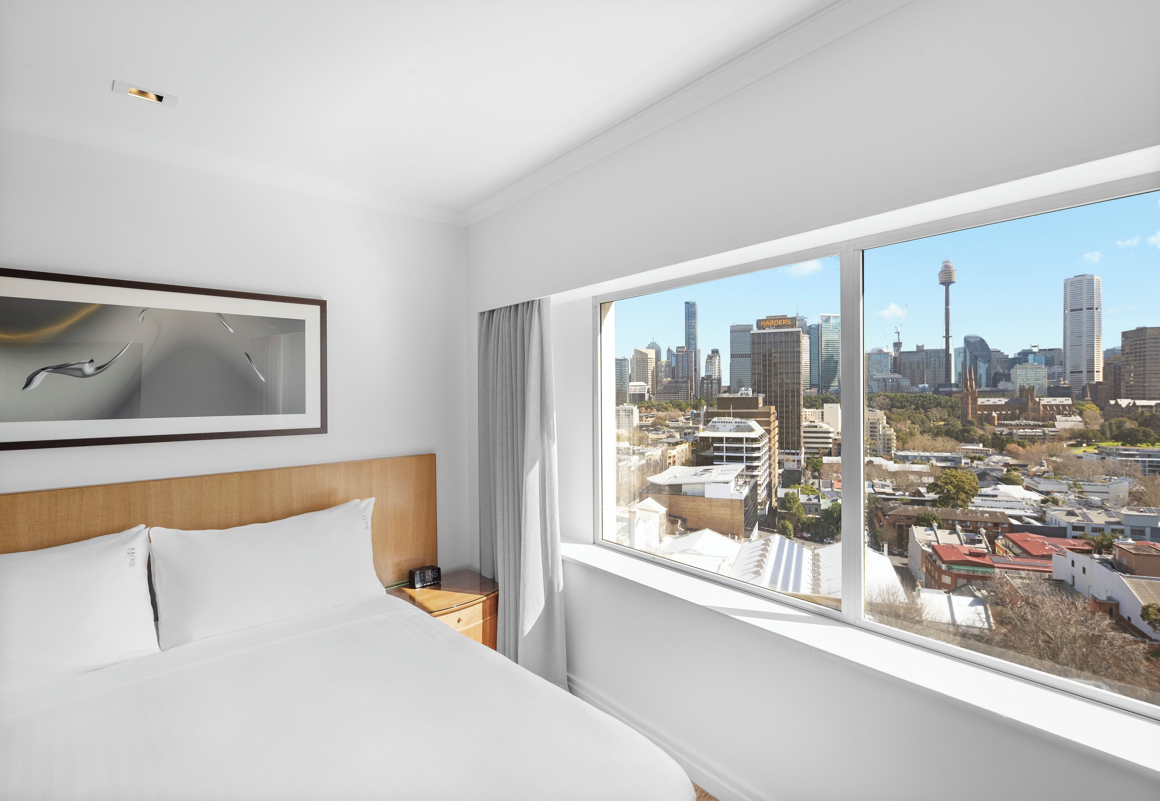 A hotel room with a large bed, a nightstand, a framed picture, and a window with a city view.