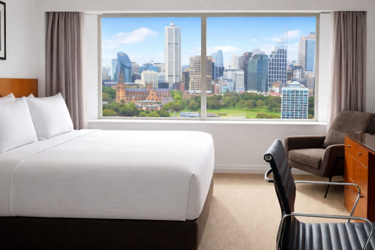 A hotel room with a large bed, a desk, a chair, and a view of a cityscape through a window.