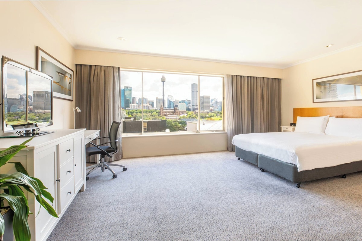 A hotel room with a bed, desk, chair, and a large window with a city view.