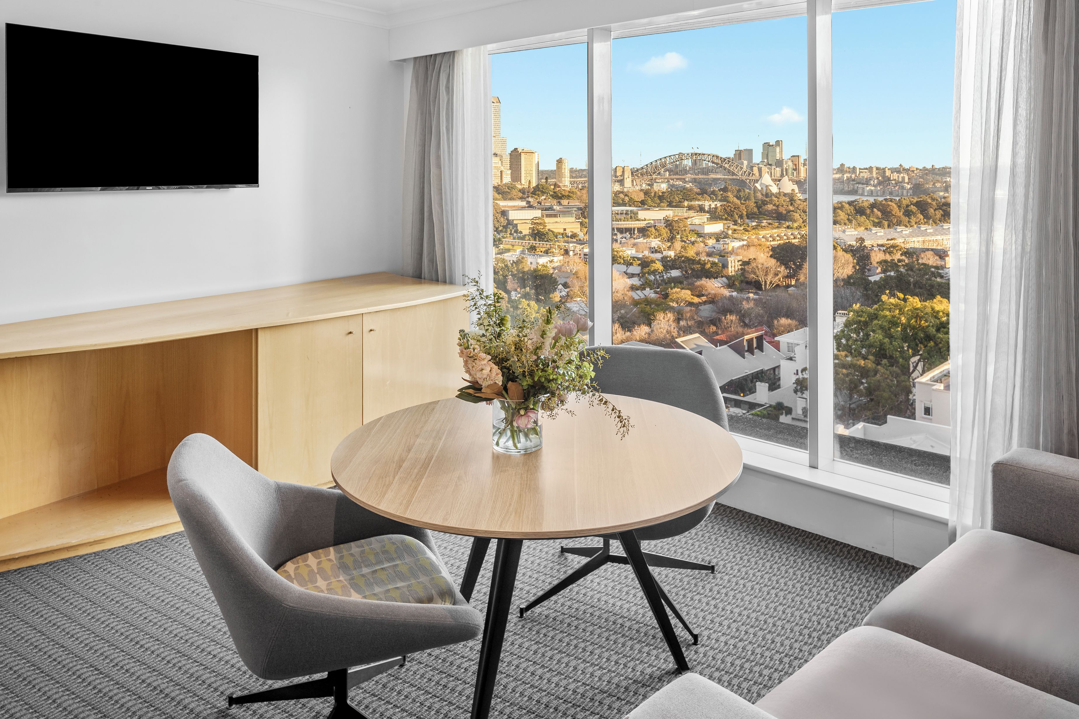 A hotel room with a round table, two chairs, a TV mounted on the wall, and a large window offering a city view.
